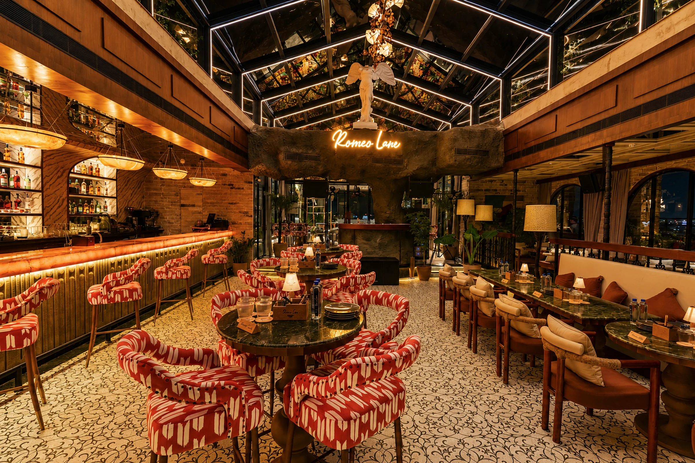 The glasshouse in the evening — coral ikat chairs under the glass roof, the Romeo Lane sign glowing on the stone wall