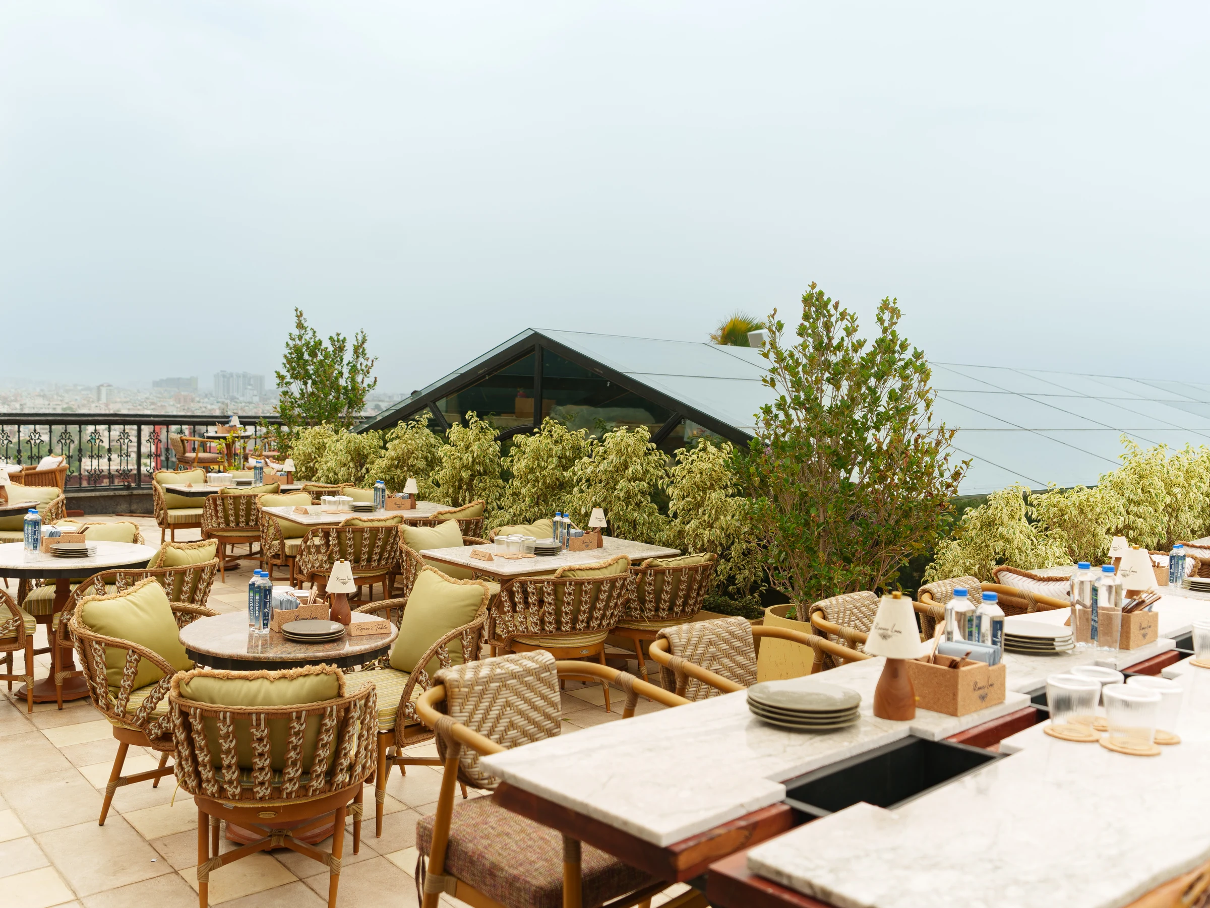The open-air terrace — cane chairs and marble tables among the plants, the city beyond